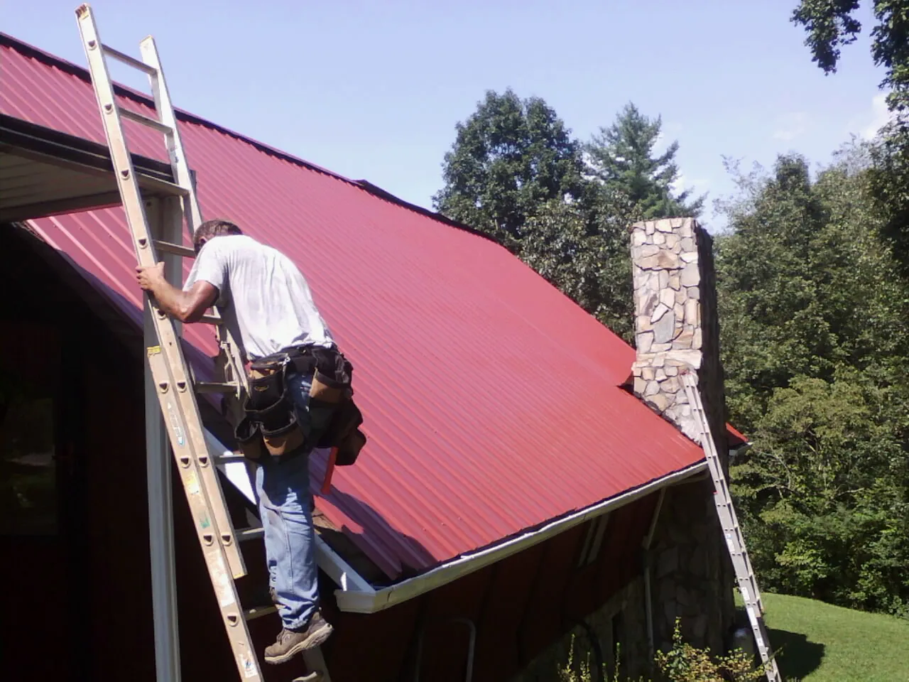 Roofer on a ladder beside a red metal roof and stone chimney — Soffit & Fascia Repair by Roofing Brothers in Winston